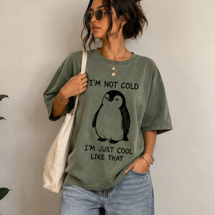A woman stands indoors wearing an oversized sage green graphic T-shirt with a penguin print and the text 'I'M NOT COLD I'M JUST COOL LIKE THAT,' paired with light blue wide-leg jeans, white sneakers, sunglasses, and a cream tote bag.